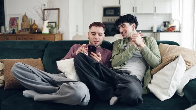 Happy male couple sitting on the living room sofa, using a smartphone. Relaxed gay partners watching content together and talking at home