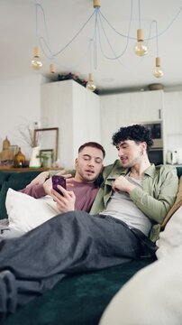 Loving gay male couple relaxing on the couch in their cozy living room, browsing the internet and shopping online with a mobile phone. Vertical