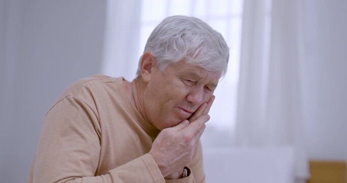 Elderly man leans forward indoors, pressing cheek and jaw with one hand while wincing and shifting head, expressing toothache, dental pain, oral health concern, and need for caring support at home.