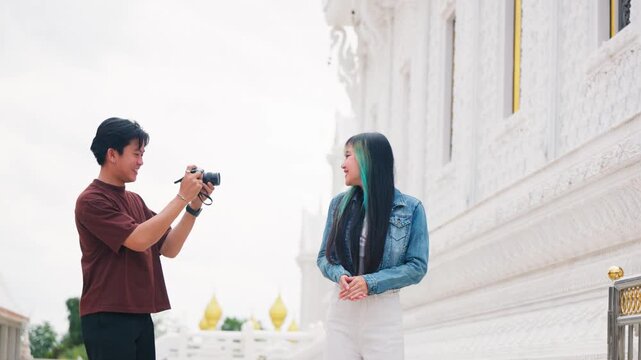 Happy Asian tourist couple exploring beautiful white temple together taking photos with camera. Travel vacation, architecture heritage tourism and cultural sightseeing lifestyle.