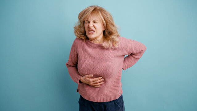Senior woman suffering from severe stomach ache and lower back pain, expressing discomfort and distress on a blue background