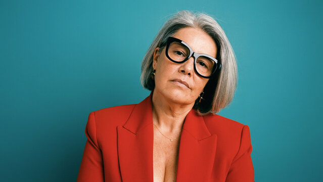 Confident mature woman wearing cat eye glasses and a red jacket, posing against a blue studio background. Professional headshot