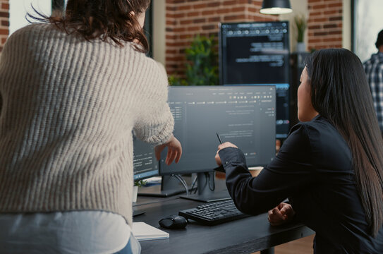 Programmer pointing pencil at computer screen with software compiling code explaining source algorithm to coworker standing next to desk around computer screens, showing developer teamwork, source.