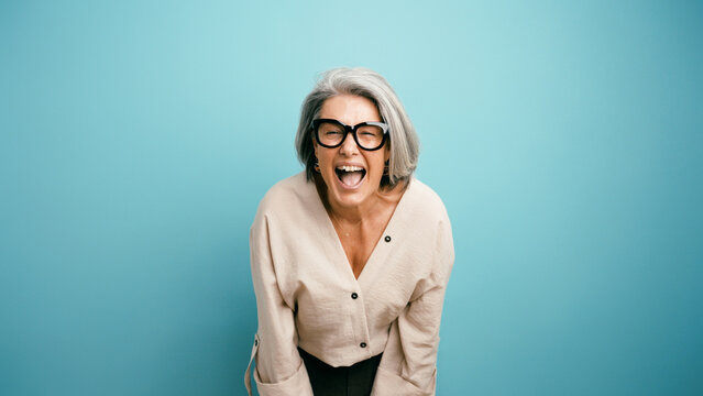 Mature woman with grey hair wearing glasses, expressing excitement and happiness while shouting on a blue background
