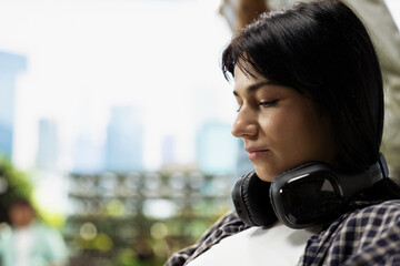 A close-up profile portrait shows a Gen Z woman with headphones around her neck sitting on campus after class and relaxing before quiet study in a bright outdoor academic environment. © DC Studio