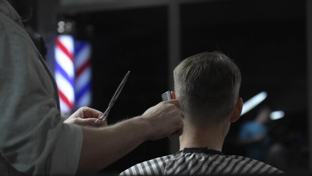 Skilled barber trimming client's hair with precision scissors, working inside vintage styled barbershop featuring traditional red white blue pole