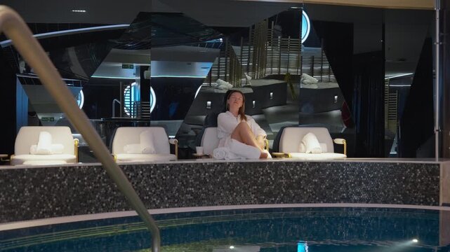 Young woman in white robe relaxes on comfortable lounge chair in upscale spa facility. Modern interior features mosaic tile pool, ambient lighting, and contemporary decor creating serene atmosphere.