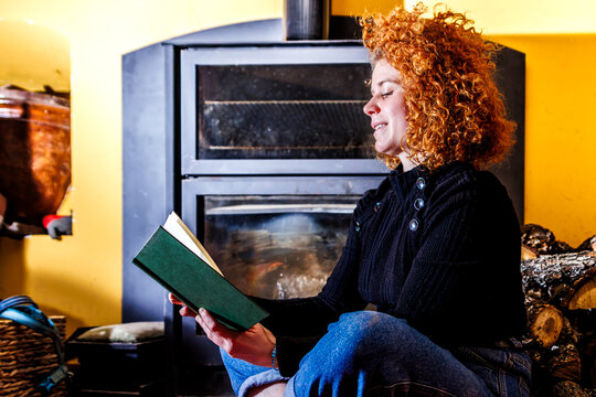 Woman with curly red hair reading book by fireplace, enjoying warm cozy home life