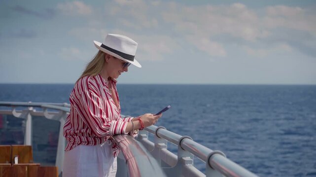 Girl enjoys cruise vacation somewhere in the ocean while checking her phone on ship deck. Blonde woman in striped shirt and white hat relaxes by railing with endless blue sea and cloudy sky behind.