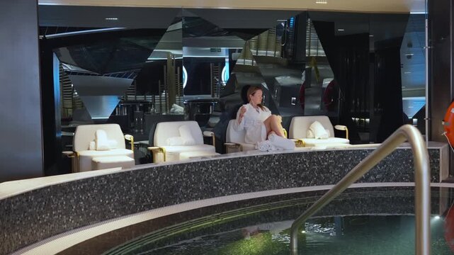 Woman in white bathrobe enjoys a relaxing moment at an upscale spa facility. She sits in a plush lounge chair beside an indoor pool with mosaic tile accents and modern mirrored decor.