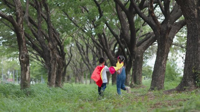 Active Asian children wearing superhero cape and mask having fun running in tree tunnel park. Kids imagination, creative future dream, and brave hero inspiration activity.