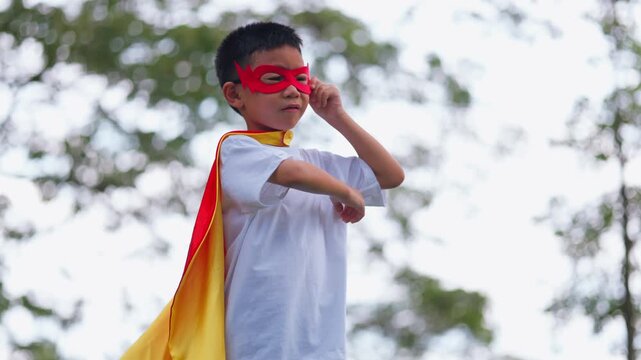 Active Asian children wearing superhero cape and mask having fun running in tree tunnel park. Kids imagination, creative future dream, and brave hero inspiration activity.