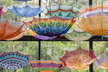 Obraz premium Artistic installation with colorful crochet umbrellas suspended indoors. Handmade lace patterns in front of windows overlooking trees. Ideal for culture and design themes.