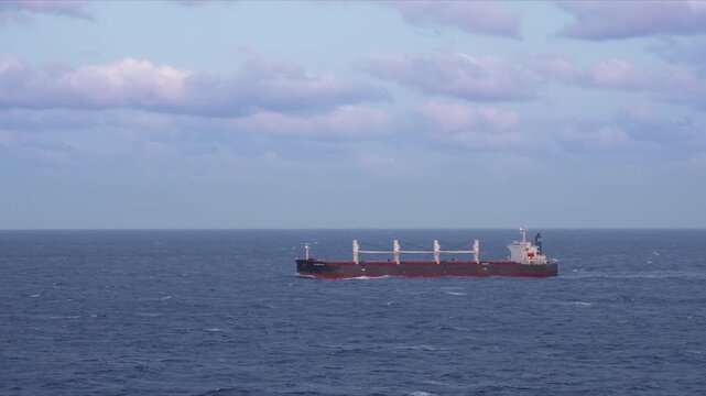 Large tanker vessel navigates through deep blue ocean waves with white crane structures visible on deck. The cargo ship crosses vast open waters under dramatic cloudy skies on an overcast day.