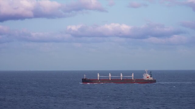 Large tanker bulk carrier vessel navigates through deep blue ocean waters with white cranes visible on deck. Ship creates wake as it travels under dramatic cloudy sky with purple and white clouds.