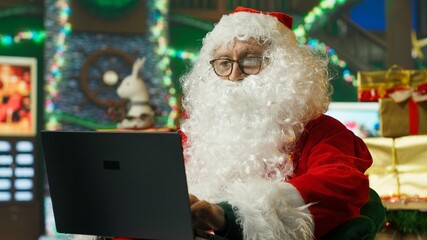 Fototapeta premium Santa Claus in a fake beard and red costume replies to children's letters on a laptop while preparing Christmas gifts in his festive workshop.