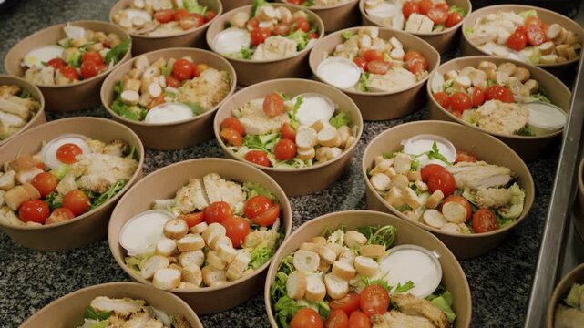Multiple bowls of fresh salad with chicken, cherry tomatoes, croutons, and dressing arranged neatly on a granite countertop in a commercial kitchen setting
