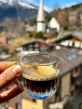 Close-up of a person holding a clear glass cup filled with fresh espresso coffee, showcasing a rich layer of crema. The background features a beautiful, soft-focus view of the traditional alpine villa