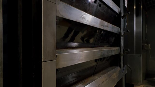 Industrial oven door with metal slats gradually opening, revealing dark interior space and reflections of surrounding equipment in a commercial kitchen environment