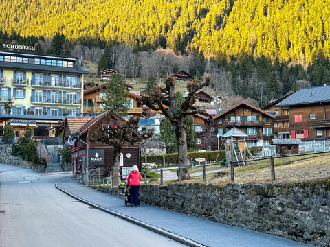 Wengen, Switzerland - 12.03.2026. A street view in the car-free village of Wengen, Switzerland, featuring a woman in a red jacket pushing a baby stroller down a paved path. The background showcases th