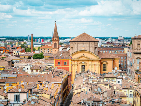 Modena cityscape captured from above, featuring historic churches, traditional terracotta rooftops, and the sprawling urban landscape under a cloudy sky