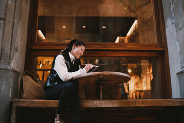 Smiling Asian freelancer working with tablet and stylus pen, creative designer browsing on mobile device at outdoor cafe, digital nomad lifestyle, remote office in evening city, tech concept. © BullRun