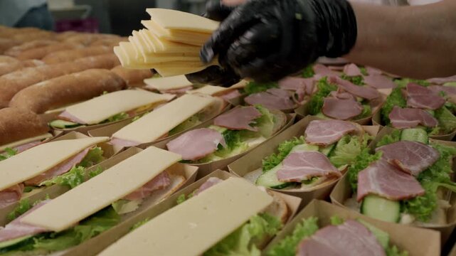 Food preparation scene showing a person assembling sandwiches with layers of lettuce, ham, and cheese in a commercial kitchen setting with fresh bread and packaging