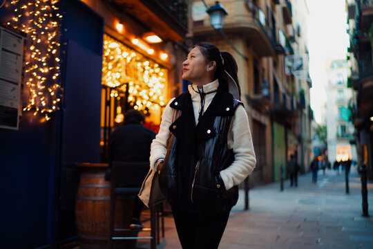 Asian woman walking on festive evening street with golden lights, trendy traveler exploring European old town during autumn vacation, authentic representation and wanderlust lifestyle.