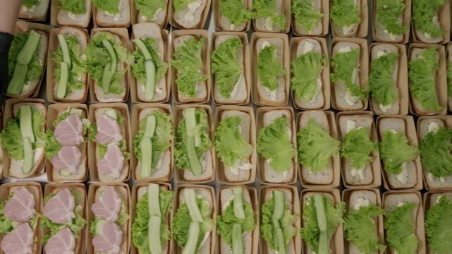 Food preparation process featuring a person assembling sandwiches with lettuce, ham, and bread in a commercial kitchen setting, showcasing organized trays of ingredients