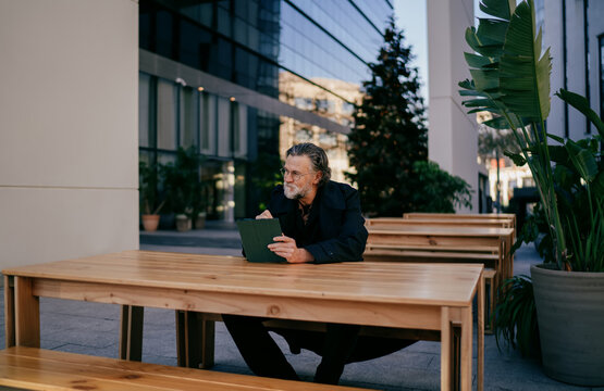 Wide shot of pensive mature businessman with tablet looking away, senior professional thinking about project at outdoor table near office building, copy space, career success, ambition.
