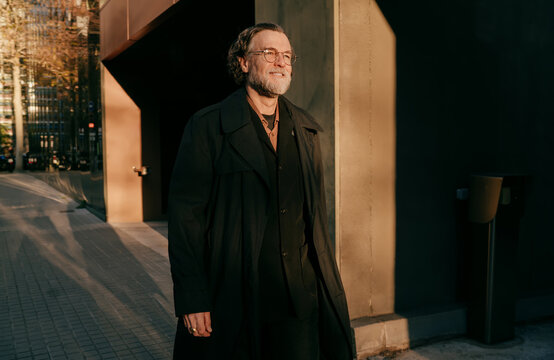 Mature confident businessman in black trench coat walking outdoors in urban business district, senior professional male manager navigating city street during golden hour with copy space.