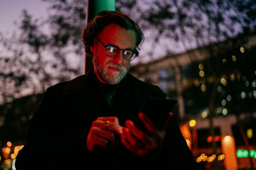 Senior male texting smartphone screen, focused man typing message mobile application night urban setting, digital nomad working remote communication, handheld device wireless technology. © BullRun