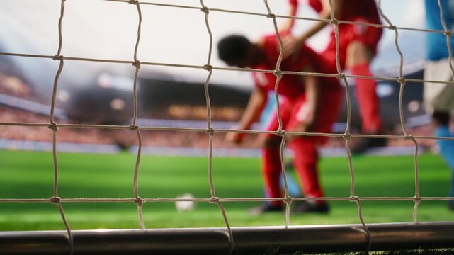Soccer player shoots ball into net across the line, with defeated goalkeeper, missed save, selective focus inside goal frame, and blurred footballers.