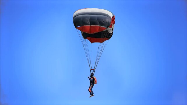A skydiver descending through the air with a deployed red, black, and white rectangular parachute/paraglider canopy against a clear blue sky background. Ideal for extreme sports, skydiving, adrenaline