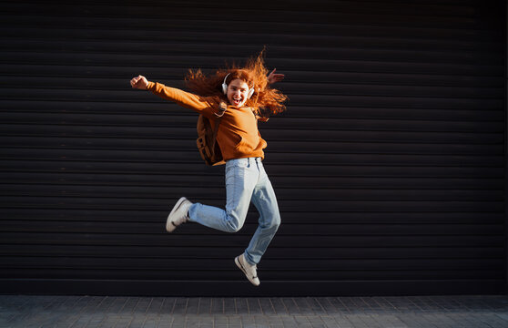 Excited redhead man with backpack jumping high on urban street, success achievement concept, active lifestyle, digital nomadic traveler, joy emotion, authentic representation, Caucasian.