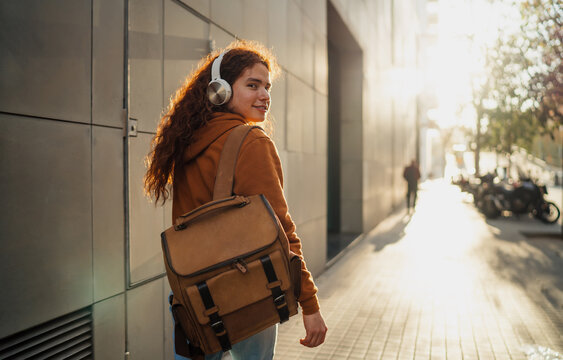 Ginger man with backpack and headphones looking over shoulder on sunny city street, digital nomadic traveler, 5G mobile connection, urban lifestyle, authentic Caucasian model, copy space.