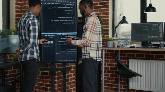 Software development team studies wall screen code during a debugging session with a digital tablet, linking error checking, while comparing programming results.