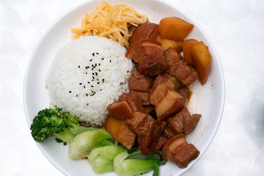 A delicious bowl of braised pork belly served with steamed white rice, bok choy, broccoli, pickled vegetables, and simmered radish. A comforting Asian meal featuring tender meat and vibrant greens.
