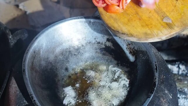 Adding chopped shallots and tomatoes to hot oil in a rustic pan for cooking over a traditional stove