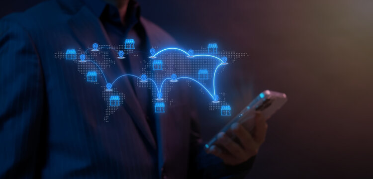 Businessman holding smartphone next to glowing world map with global retail shop stores and location pins icon. Dark neon blue lighting, international franchise expansion and shop placement concept.