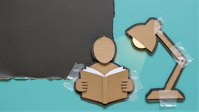 Cardboard cutout of person reading book with desk lamp, taped to teal background.