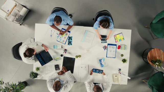 Specialists and medics discuss therapy options from an overhead angle in a hospital office, where clinical information and treatment notes help them support care planning inside a medical team.
