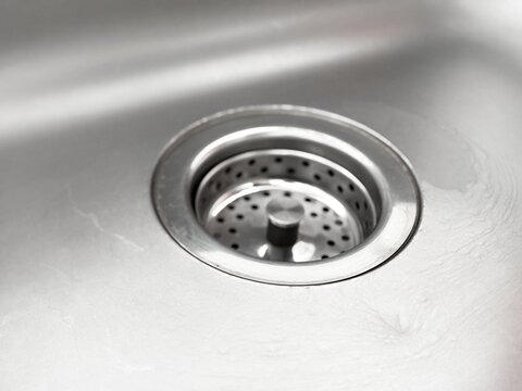Closeup view of drain with circular metal strainer with holes in kitchen sink for washing dishes. Sink surface appears smooth and shiny, details of metal. Details of interior