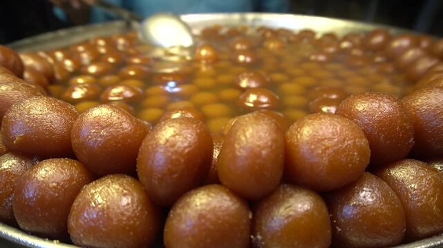 A large metal tray filled with sweet brown gulab jamun balls completely soaked in sugary syrup at a sweet shop