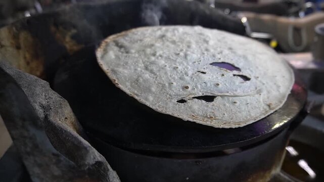 A round white flatbread baking chapati on a convex metal tawa placed over an open wood fire in a traditional kitchen