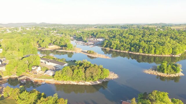 Aerial perspective of a serene river scene in Johnson City, Tennessee, featuring vibrant green trees, rocky banks, and still waters mirroring the blue sky, with structures and boats lining the shore