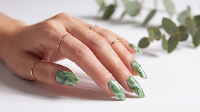 Hand wearing thin gold rings showcases long, tapered nails with a striking green marble pattern, complemented by delicate eucalyptus foliage on a clean, light-colored surface for a fresh look.
