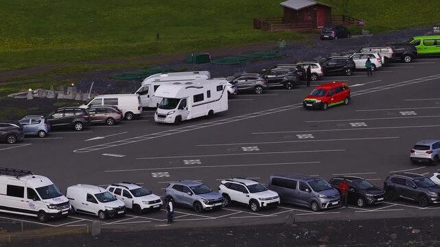 Aerial view of the Reynisfjara area near Vik, Iceland, with rows of cars, campervans, a tour bus, and a service van moving under overcast light by the black sand shore.