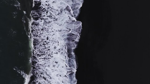 Aerial top down view of Vik black sand shoreline in Iceland as waves break and recede, white surf forming lacy patterns under overcast light, no people present.