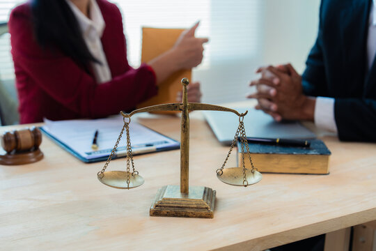 Close up of justice scales and judge gavel on wooden desk with cash bundle and lawyer consulting client about financial lawsuit in blurred background.